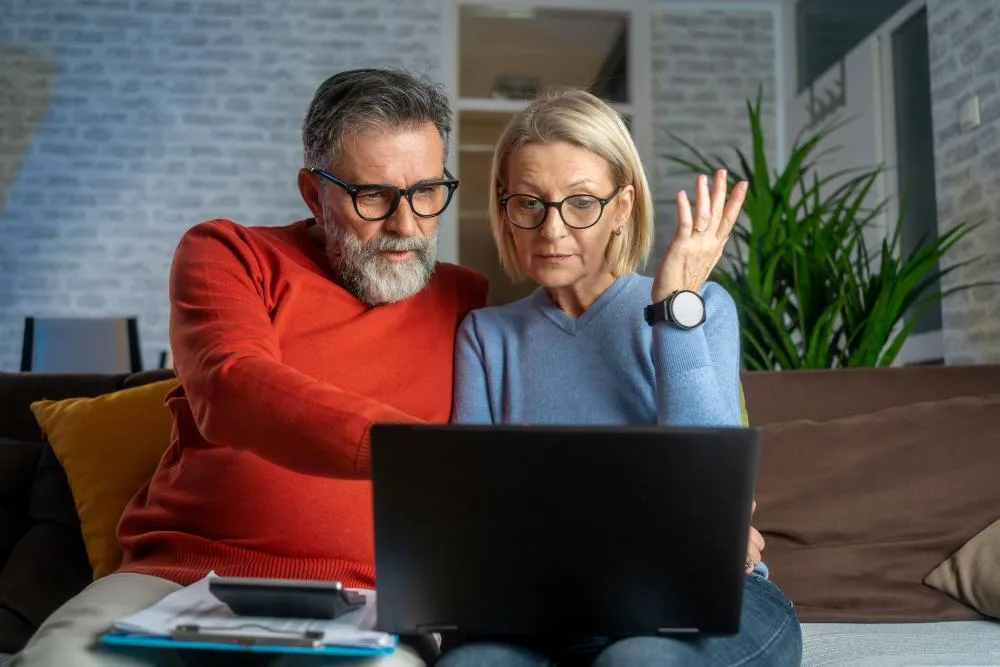 retirement-age couple sits on couch looking at laptop