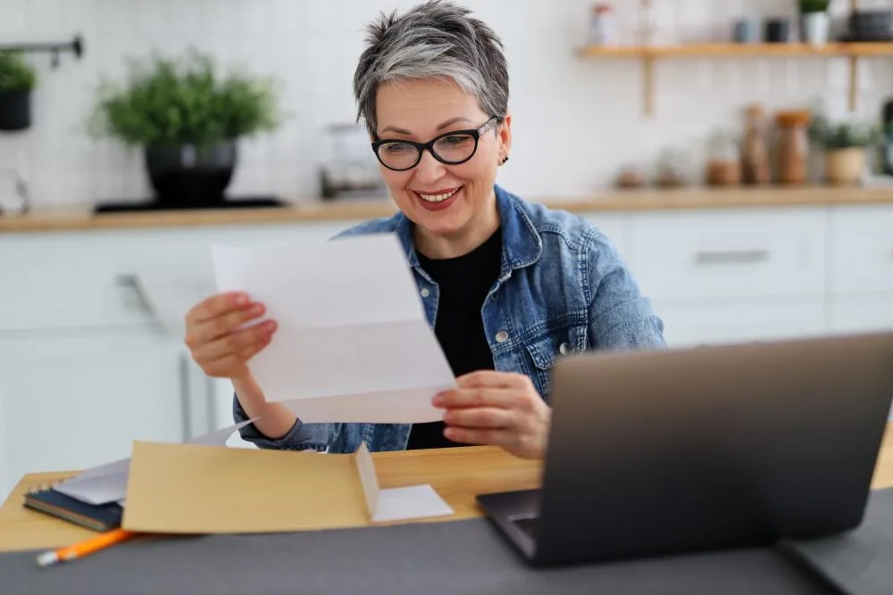 woman with short gray hair and glasses smiles looking at a piece of paper