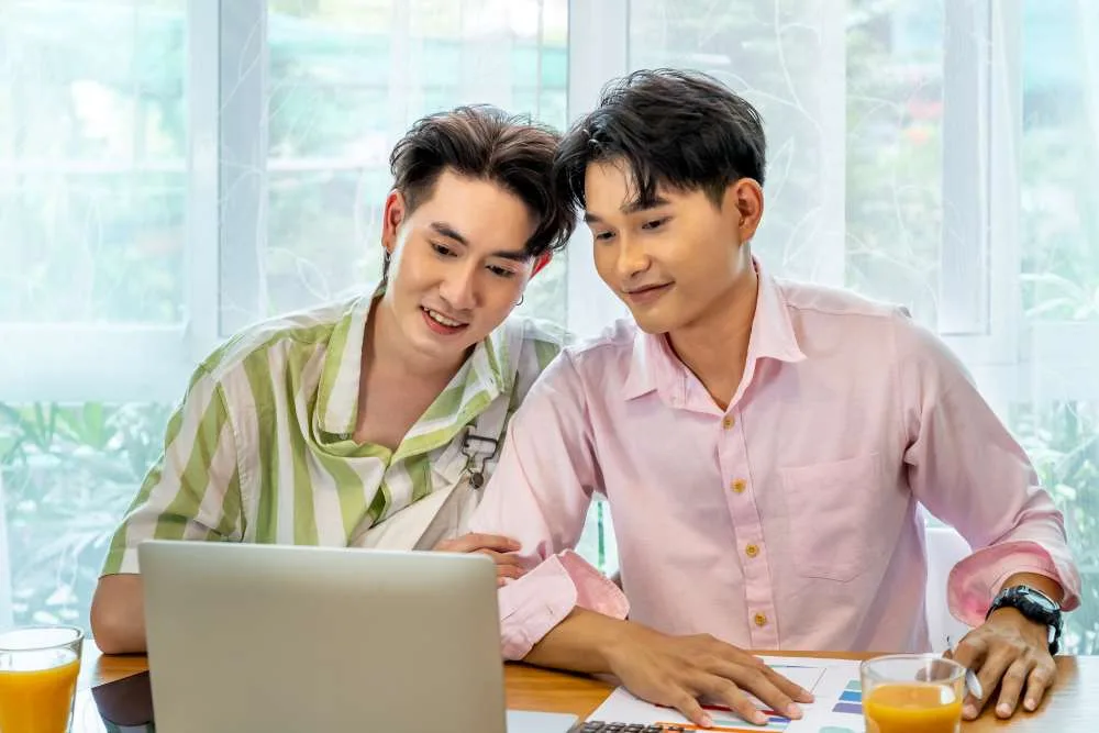 Two men, one in pink shirt, one in green striped shirt, sit looking at laptop