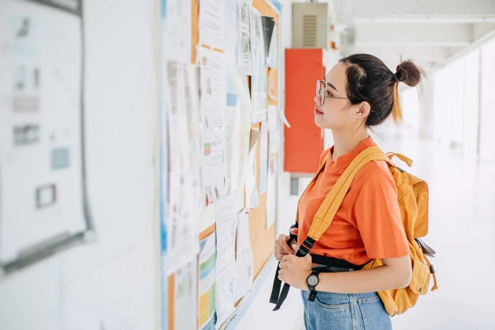 Young woman wearing backpack looks at board covered in job flyers