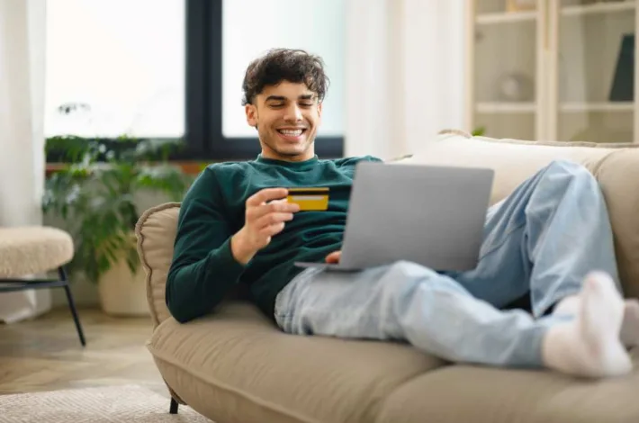 Young man sits on couch with laptop and holding credit card