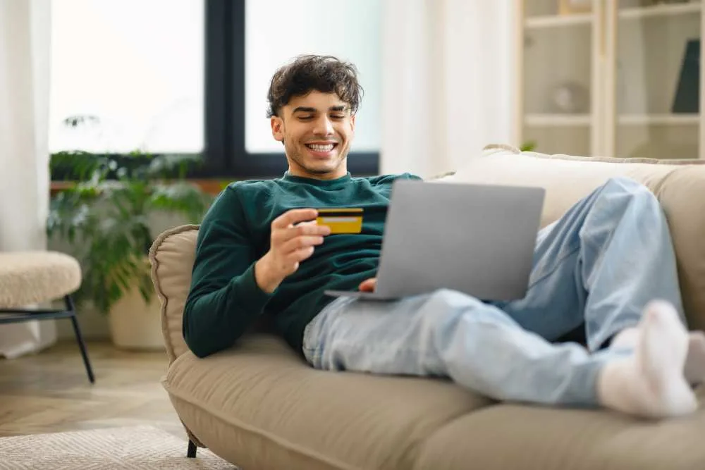 Young man sits on couch with laptop and holding credit card