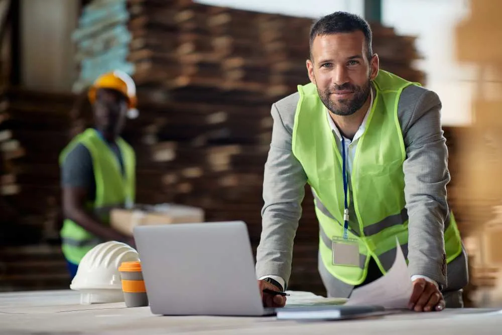 Male business owner in suit and safety vest works on laptop in a warehouse