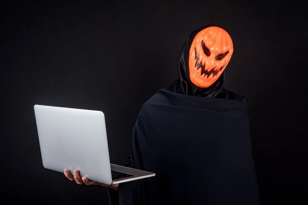 Person wearing scary pumpkin Halloween mask holds a laptop computer