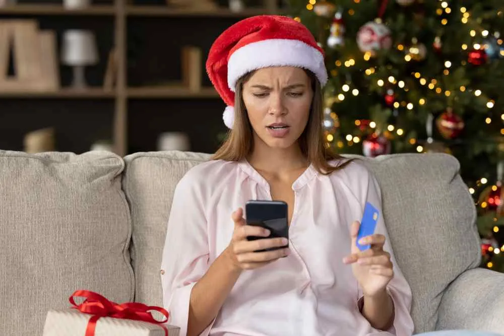 Woman in Santa hat sits in front of Christmas tree looking annoyed at her phone and credit card