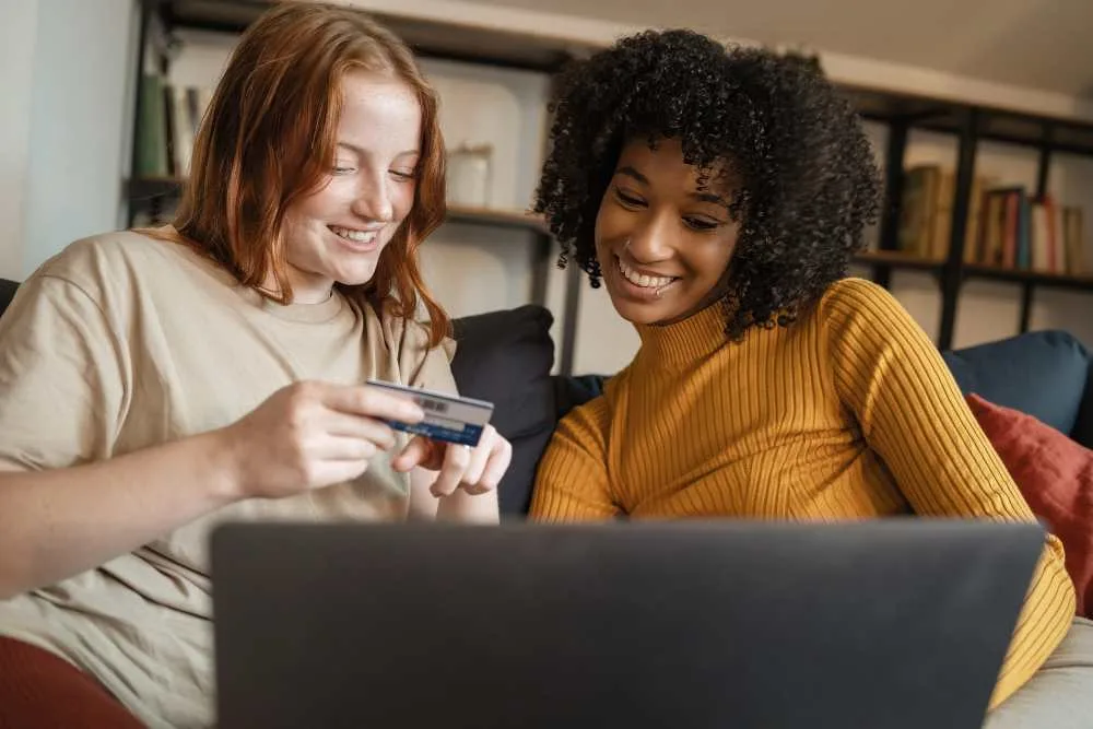 Two women sitting on couch with credit card looking at laptop