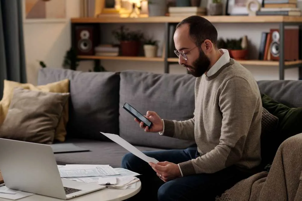 Man with glasses holds phone, sits on couch doing his taxes