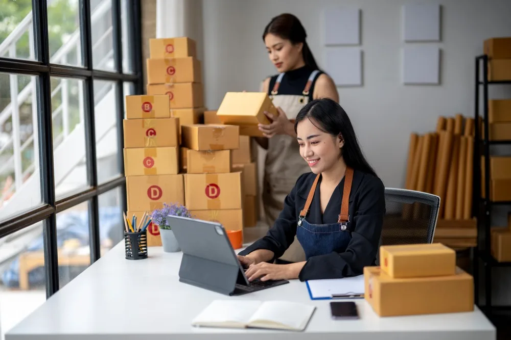 Two young businesswomen working with packages in a warehouse using digital tablet